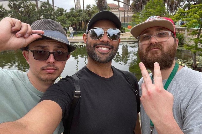 Drie mensen maken een selfie bij een waterpartij, met een brug en palmbomen op de achtergrond. 2