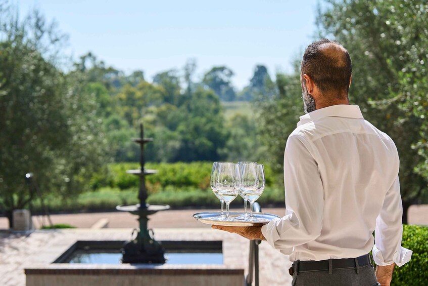 A man carrying a tray with wine glasses and a water feature in the background. 3
