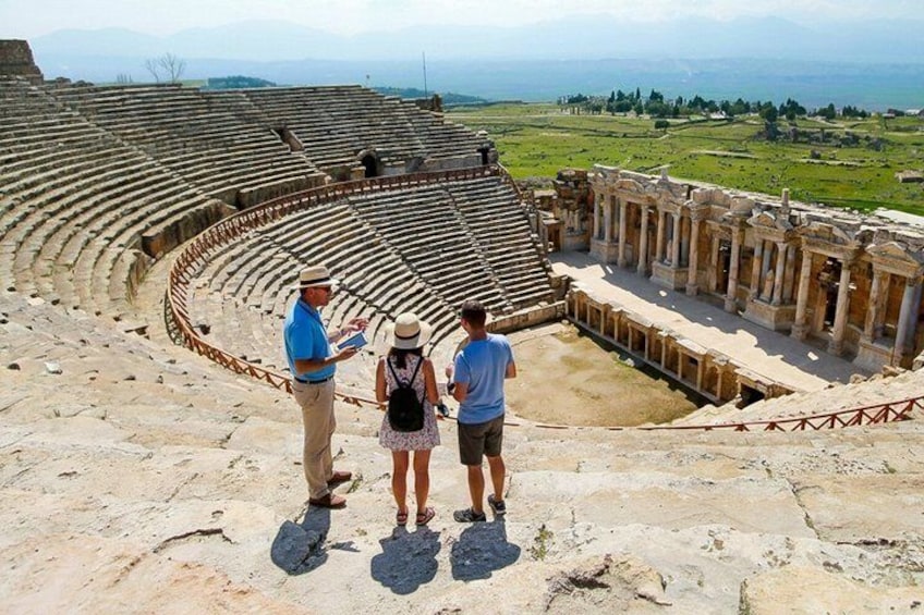 An ancient amphitheater with stone seating and a central stage, surrounded by ruins and a hilly landscape. 4