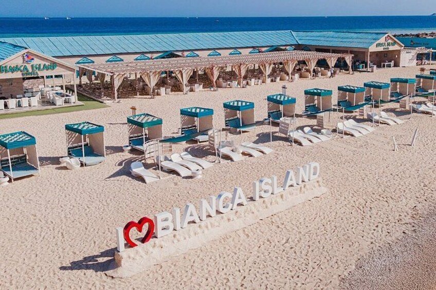 En strand med solstolar och parasoller, en skylt med texten ”BIANCA ISLAND”, och en byggnad med ett blått tak. 4