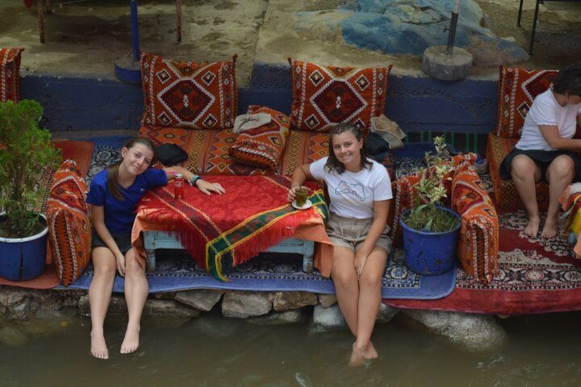 Two people sitting on a bench by a water body, with colorful cushions and potted plants around them. 1