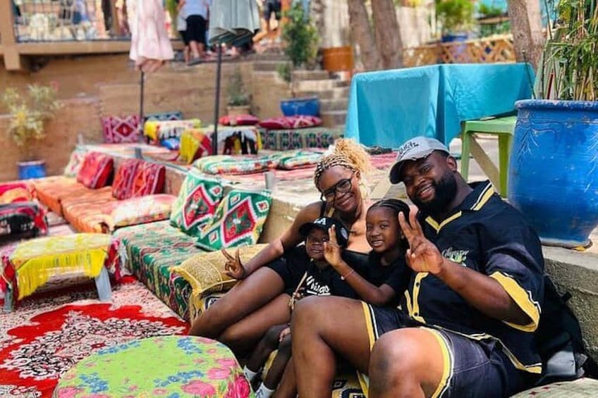 A group of people sitting on colorful textiles outdoors, with a market in the background. 1