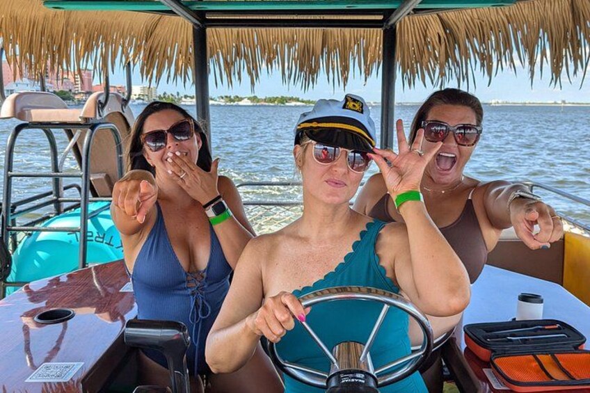 Three people on a boat with a steering wheel, a table, and a thatched roof. 1