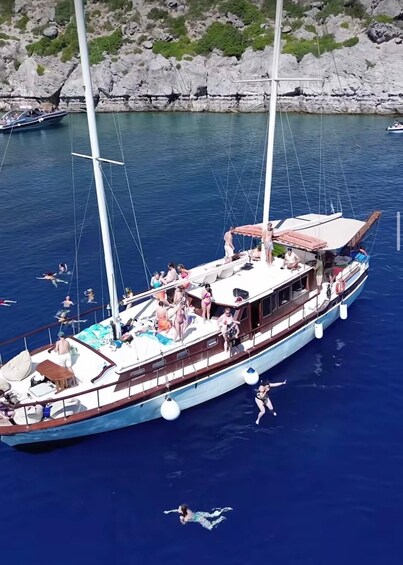A large sailboat docked at a rocky coastline with people swimming and lounging on the deck. 5