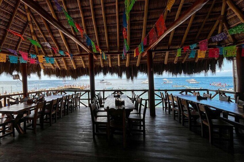 An empty beachside restaurant with wooden tables and chairs, colorful flags, and a view of the ocean. 3