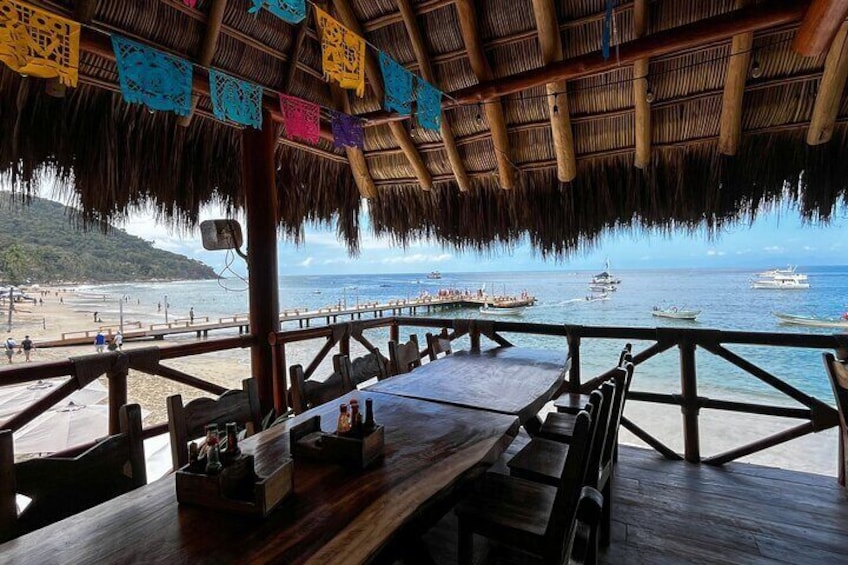 A wooden deck overlooking a beach with a thatched roof, wooden chairs, and a table with bottles. 2