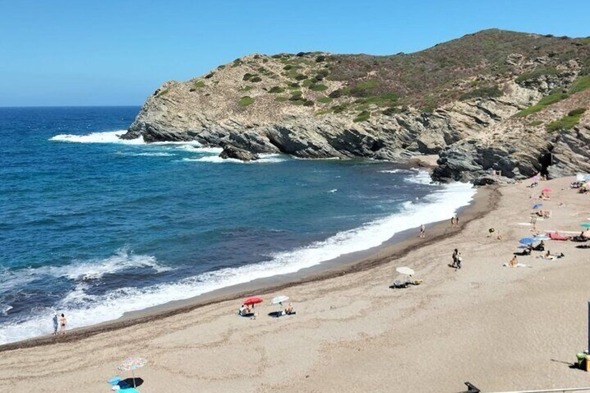 Ein Strand mit felsigen Klippen, klarem, blauem Wasser und sonnenbadenden Menschen. 5