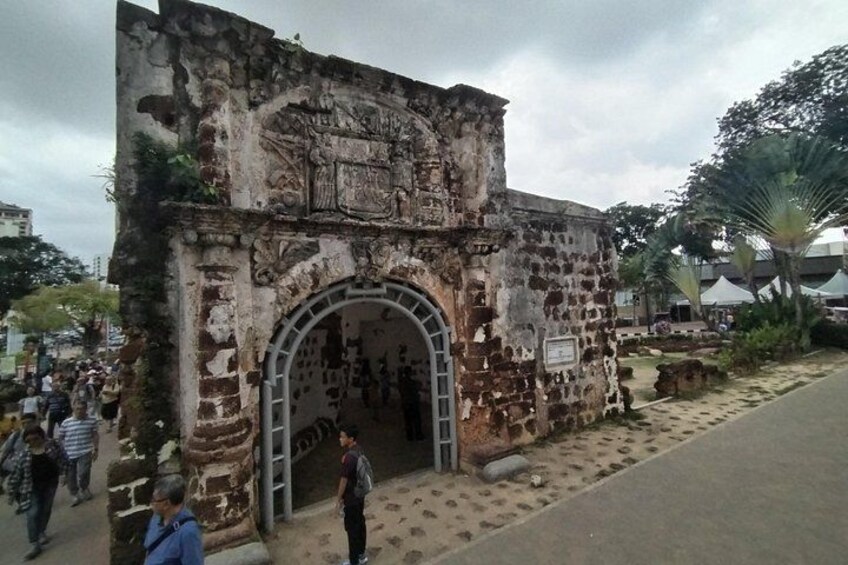 An old stone building with a large arched entrance, surrounded by people and palm trees. 1