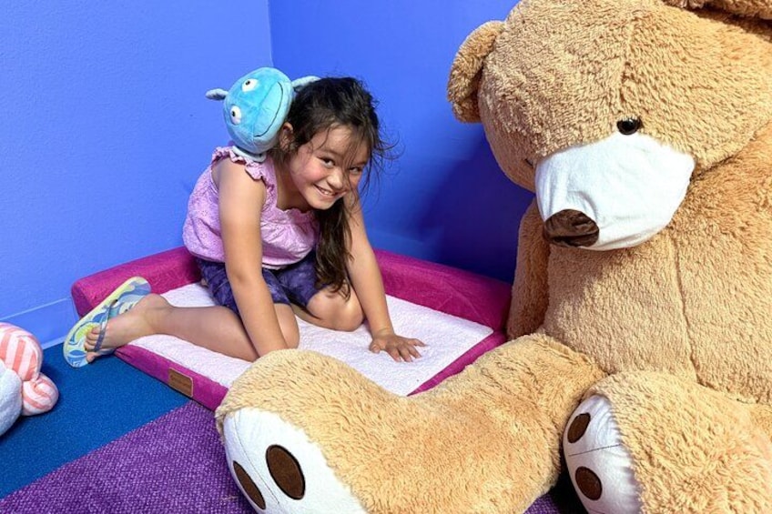 Una niña sentada sobre una alfombra rosa con un osito de peluche grande a su lado. 2