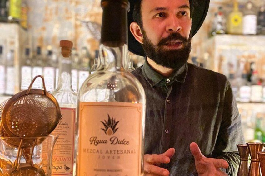 A person standing behind a counter with bottles labeled 'Rigua Dulce Mezcal Artesanal Joven' and a strainer. 4
