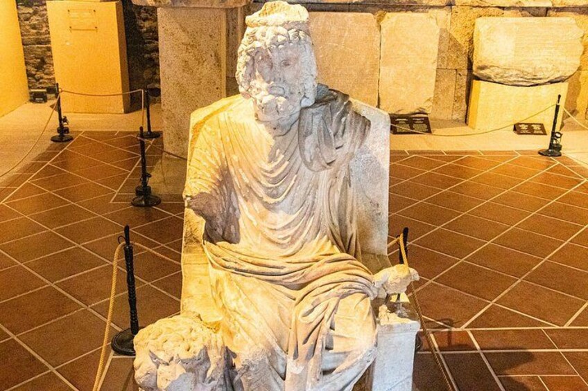 A marble statue of a seated figure with draped clothing, placed on a tiled floor with ropes and stanchions for protection. 2