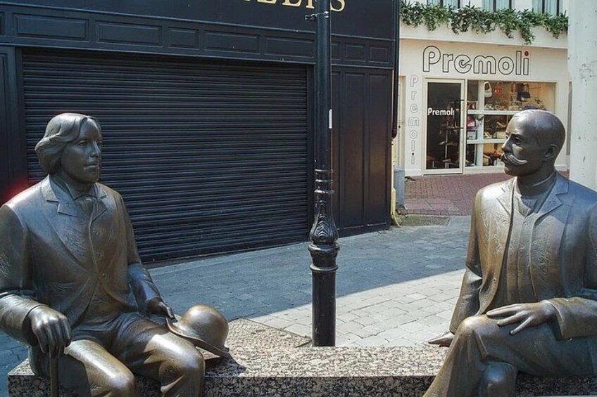 Two bronze statues of men sitting on a bench, a street lamp, and a storefront with the name 'Premoli'. 2