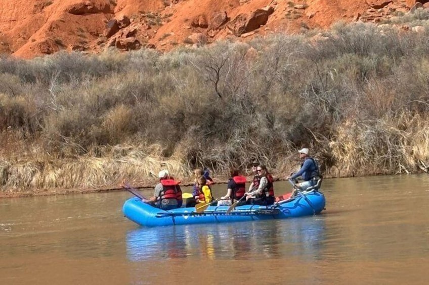 A group of people in a blue inflatable raft on a river with red rock formations in the background. 5