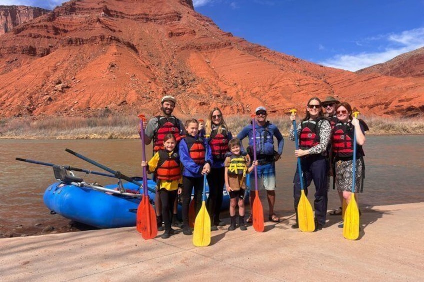 A group of people in a blue raft on a river with red rock formations in the background. 1