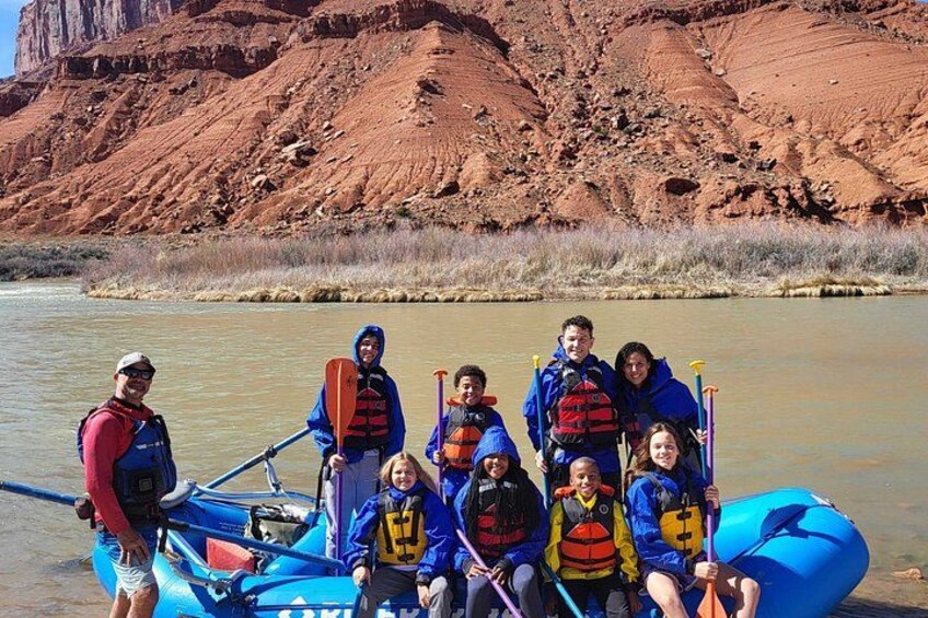 A group of people in a blue raft on a river with red rock formations in the background. 4