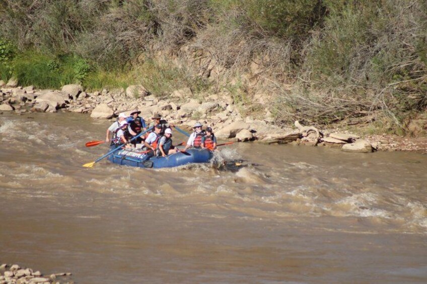 A group of people in a blue raft navigating a river with rocks and vegetation along the banks. 2