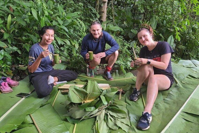 Three people sitting on a mat made of large leaves, engaged in an outdoor activity. 3