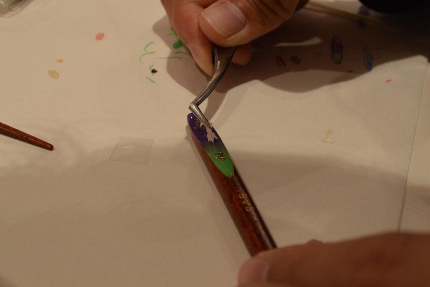 Una mano che impugna un pennello con manico in legno e rivestimento metallico, applicando vernice su una superficie bianca con un piccolo contorno quadrato. 2
