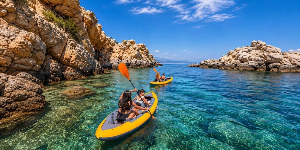 Two people kayaking on a clear blue sea with rocky cliffs. 1