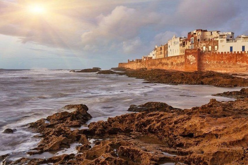 A coastal town with a fortified wall, rocky shoreline, and a sunset. 5
