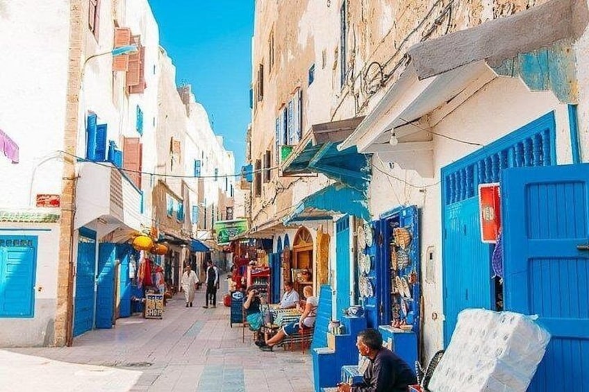 A narrow street with white buildings and blue doors, shops, and people sitting outside. 1
