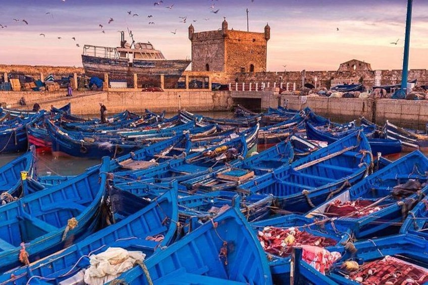 A harbor with numerous blue boats docked, a large stone tower, and a clear sky. 2