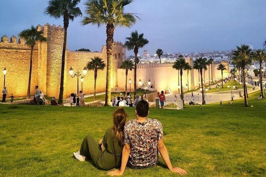 A couple sitting on grass in front of a historic fort with palm trees and a cityscape in the background. 1