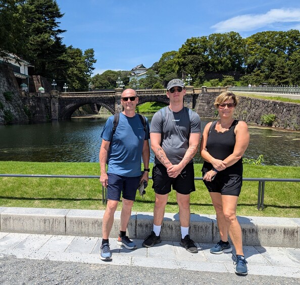 Three people standing by a river with a stone bridge and greenery in the background. 3