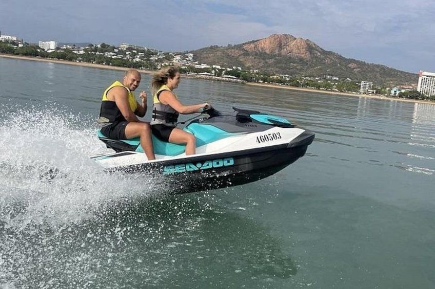 Un jet ski avec deux personnes à bord, de l’eau qui éclabousse tout autour, et un paysage montagneux en arrière-plan. 3