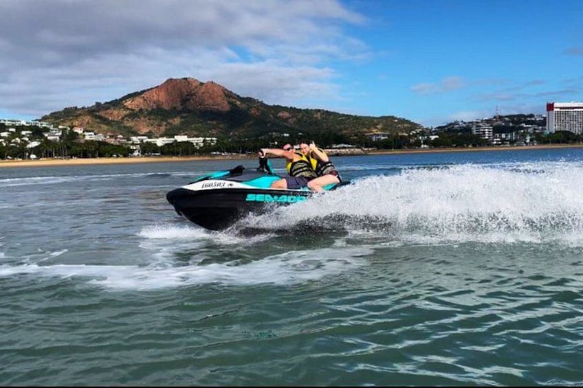 Un jet ski avec deux personnes à bord, de l’eau qui éclabousse tout autour, une montagne en arrière-plan et une plage avec des bâtiments. 1