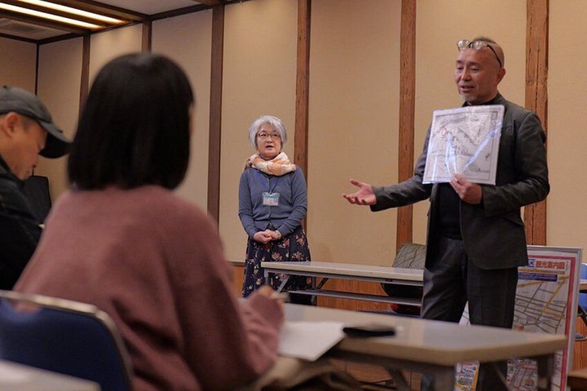 A person presenting a map to an audience in a room with wooden paneling. 2