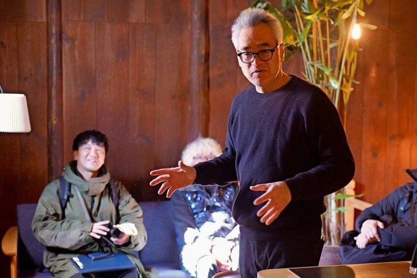 A man gesturing while standing in a room with wooden walls and a plant in the background. 1