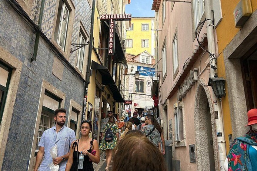 A narrow street with buildings, a restaurant sign, and people walking. 1