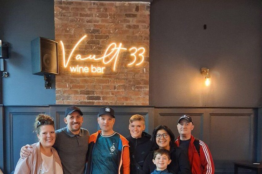 A group of people standing in front of a brick wall with a neon sign that reads 'Vault 33 wine bar'. 5