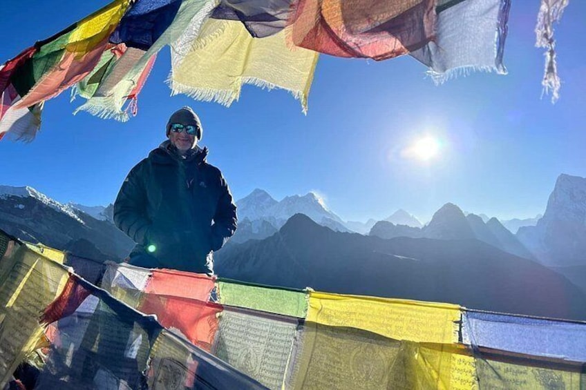 Une personne se tient sur une crête montagneuse, des drapeaux de prières flottent autour d’elle, et on aperçoit des montagnes et un ciel dégagé en arrière-plan. 4
