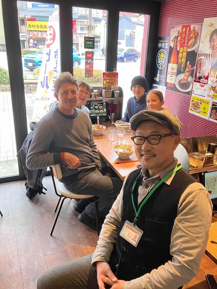 A group of people seated at a table inside a restaurant with a menu board and a window view of the street outside. 5