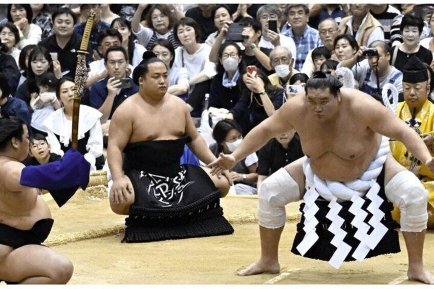 Two sumo wrestlers in traditional attire, one seated and one standing, with a crowd in the background. 3