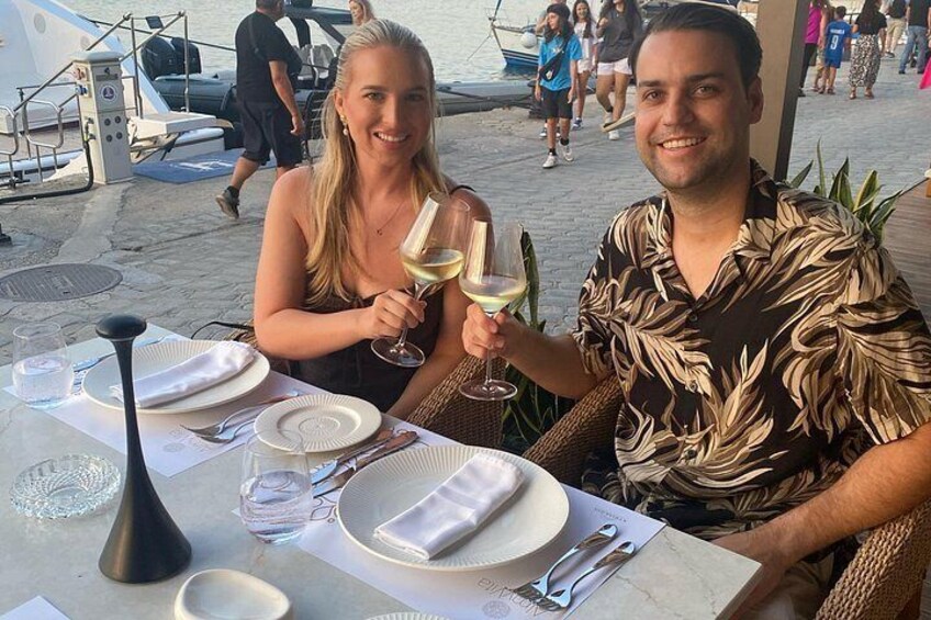 Two people at a table with a glass of white wine, a boat in the background, and a beach setting. 1