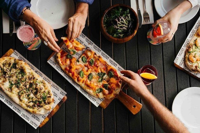 A person holding a pizza on a wooden board, with another pizza on the table, a bowl of salad, and drinks. 3
