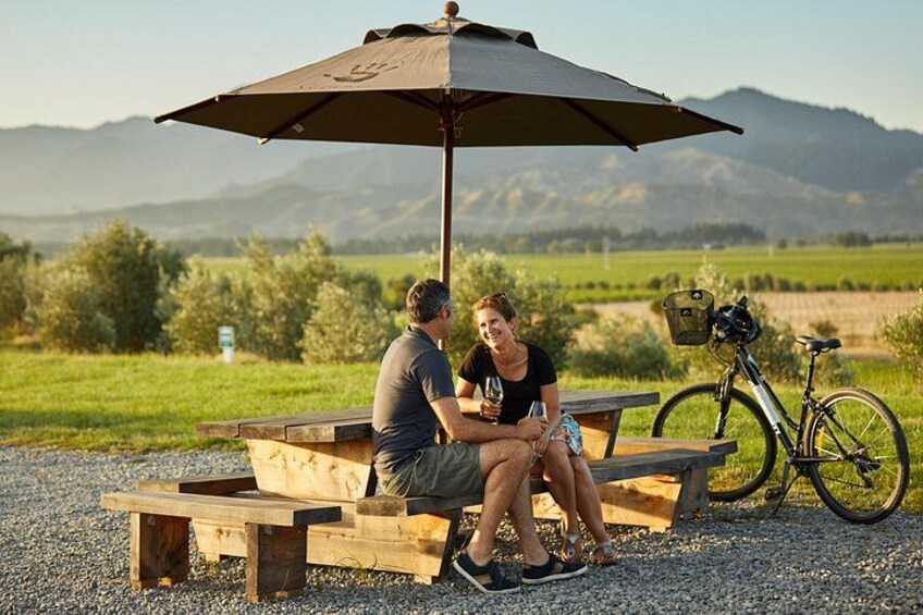 Een stel zit op een houten bank onder een grote parasol, met een fiets die er geparkeerd staat. 5