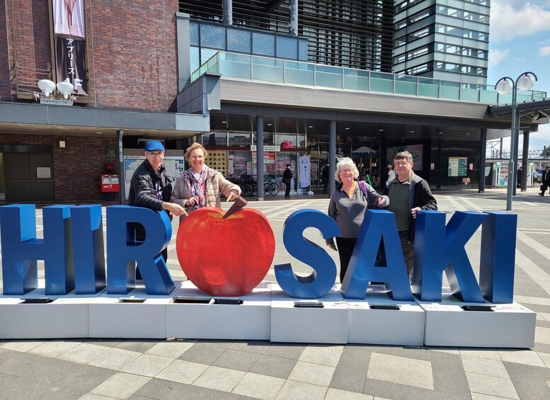 A sign with the letters 'HIR' and 'SAKI' in blue, a red apple sculpture, and people standing in front. 1