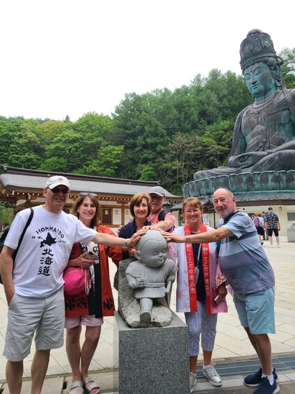 A group of people posing in front of a large statue and a smaller stone figure, with traditional Japanese architecture in the background. 3
