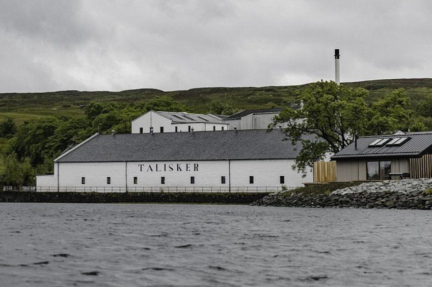 A white building with 'TALISKER' written on it, a body of water in the foreground, and a hill with greenery in the background. 3
