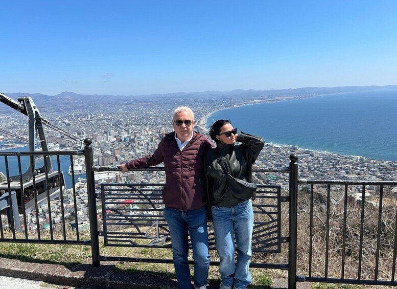 Two people standing by a railing overlooking a coastal cityscape with mountains in the distance. 3