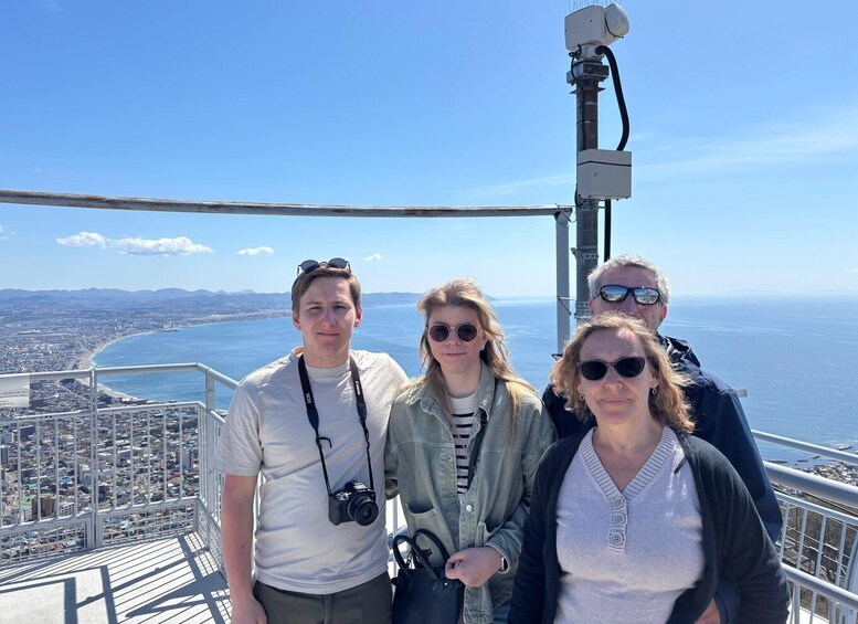 A group of people on a viewing platform overlooking a coastal city with a clear view of the ocean and distant mountains. 2