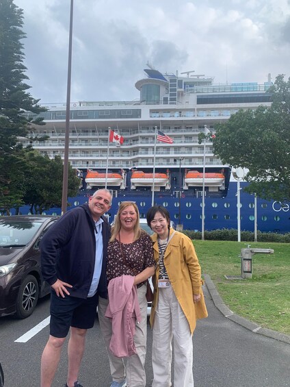 Three people standing in front of a cruise ship with Canadian and American flags, a parking lot, and a building with a blue facade. 2