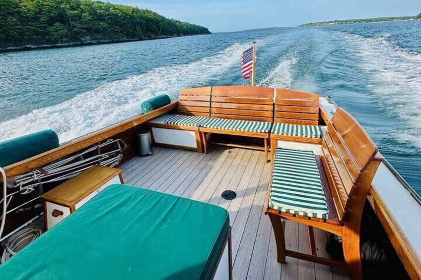 A boat deck with wooden benches and a table, a flag, and a view of the water and land. 2