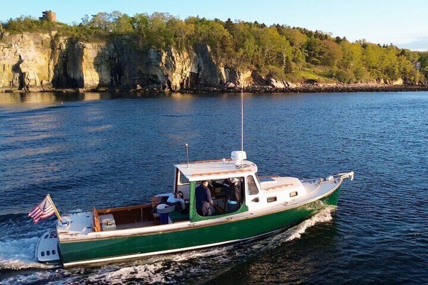 A green boat with an American flag on the bow, moving on calm water with a rocky shoreline and trees in the background. 3