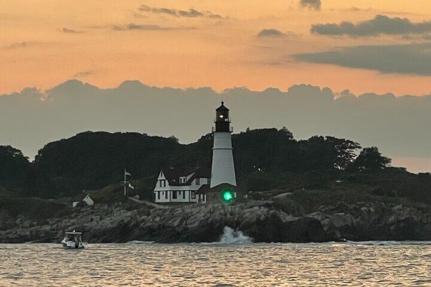 A lighthouse with a green light, a house, and a boat on a rocky shore at sunset. 5