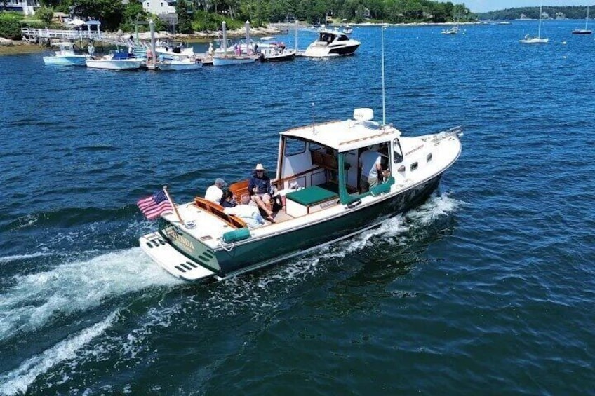 A green and white boat with an American flag on the bow, moving on a body of water with other boats in the background. 1